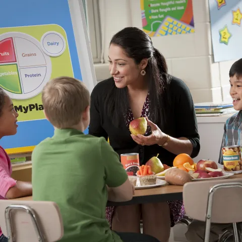 A teacher sits at a table with elementary-school aged students and talks with them about healthy eating, while holding a piece of fruit in her hands. The MyPlate graphic is visible on a poster in the background.
