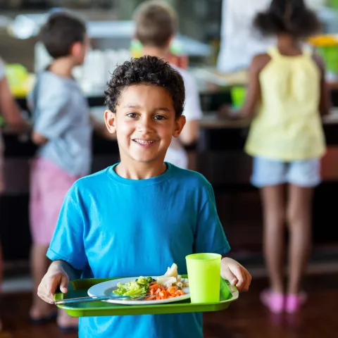 Young boy in blue shirt standing in a school cafeteria holds a green reusable tray with a healthy school meal.