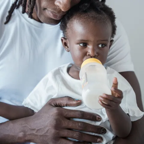 Image on the left of a young girl drinking milk out of a bottle while being held by her father. Photo by Sasha Kim.