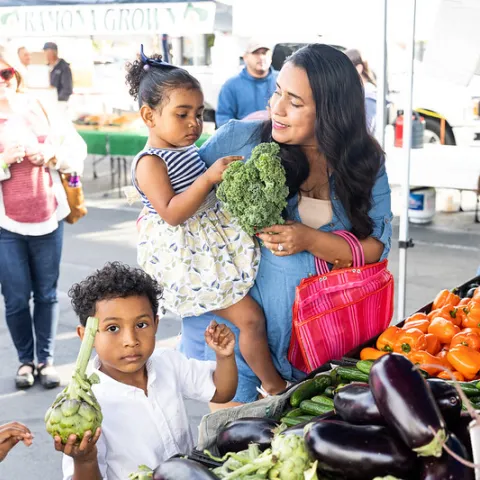 A mother holds her daughter in her arms while her son stands next to her holding fresh vegetables he selected at a farmers' market booth.