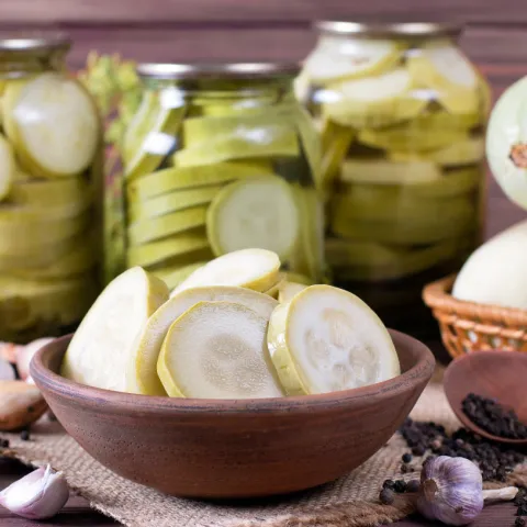 A rustic display of preserved zucchini. Several glass jars in the background are filled with neatly sliced rounds of pale green zucchini. In the foreground, a wooden bowl holds additional slices, and whole zucchini sit nearby in a woven basket. Garlic, small onions, peppercorns, and a wooden spoon are scattered around the burlap surface, giving the scene a natural, homemade food preparation feel.