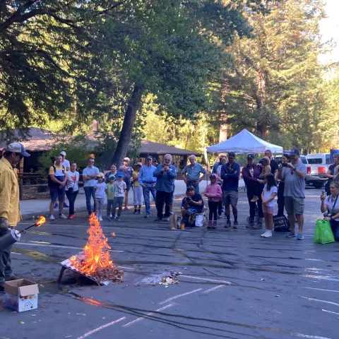 A crowd of kids and adults watch a fire expert light a fire on an inclined board 