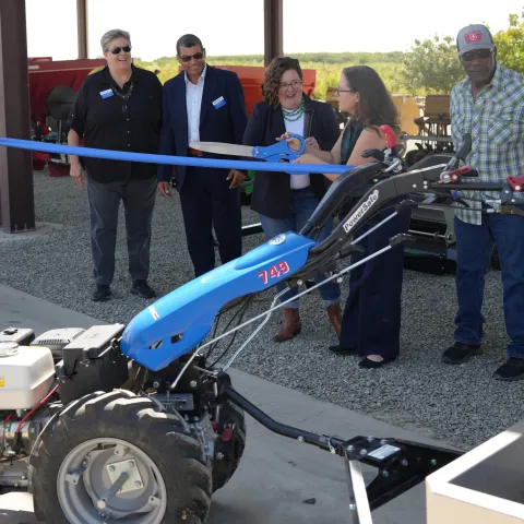 A group of people prepare to cut a ribbon strung between two pieces of farm equipment