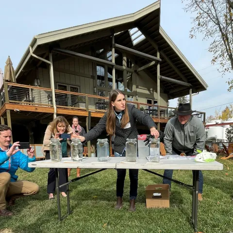 Hope, speaking to people gathered around a table, gestures to five glass jars with soil clods floating in water