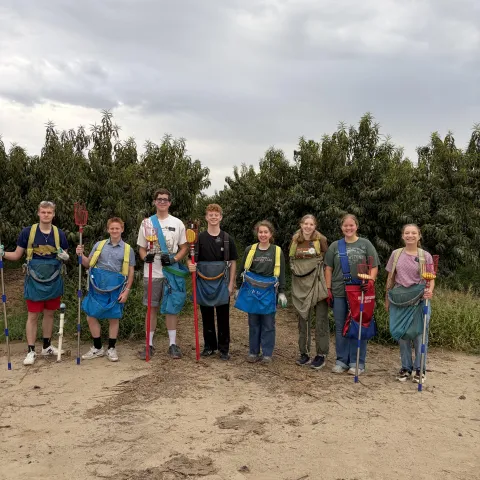Eight people wearing fruit picking bags and holding harvest tools pose beside orchard.