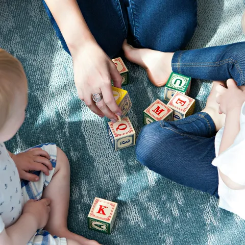 Two toddlers and a caretaker play with wooden lettered blocks on the floor