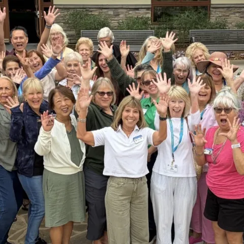 A large group of about 35 people stands outdoors on a stone patio, waving and raising their arms in the air, smiling, in front of a stucco building with wood-trimmed windows and surrounded by trees and shrubs.