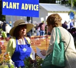 A woman in a straw hat and blue apron provides plant advice at a garden fair, while speaking with another woman in a light sweater.