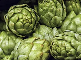 A cluster of fresh artichokes varying in size and shape, with textured, green leaves and some hints of browning at the tips. The background is dark.