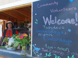 Smiling volunteer at a farm stand booth beside a chalkboard sign welcoming community volunteers Tuesdays–Saturdays, with fresh-cut flowers in the foreground.