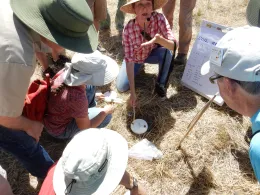 An instructor with a microphone demonstrates soil respiration testing to a group outdoors, with a chart showing soil readings nearby.