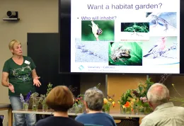 A woman in a green "Going Native Garden Tour" shirt presents a slide titled "Want a habitat garden?" showing wildlife photos, to a small seated audience.