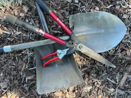 A collection of garden tools, including a metallic shovel, flat trowel, long-handled tools, and pruning shears, arranged on mulch.