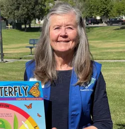 Master Gardener Denise smiling and wearing a blue vest while holding the life cycle of butterfly (complete metamorphosis).