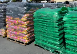 Pallets of brightly colored soil mix bags labeled "Raised Bed" in yellow and pink, alongside green bags labeled "Garden Soil," stacked in a storage area.