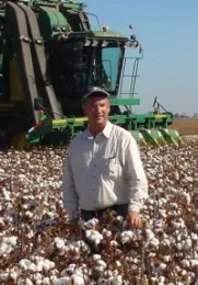 Munk stands in a field of cotton ready for harvest, with a green cotton harvester in the