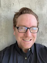 Eliot Freutel wearing glasses and a black shirt, and a gray background