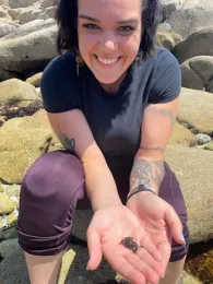 A woman in a dark shirt and magenta pants, with rocks in the background