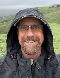 A man with glasses and a dark jacket smiling, with green hills in the background