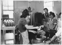 Black and white photo of the women demonstrating canning process in front of group of women.