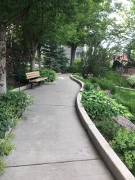 A concrete pathway curves through a lush garden with trees and a bench. Purple allium flowers are visible among the green foliage.