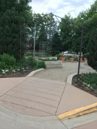 A metal garden archway frames raised flower beds and potted plants in a garden setting. Wide, sloped, paved, textured walkways with curbs lead through the garden, with lush green trees and a cloudy sky in the background.