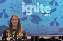 A 4-H youth stands in front of an Ignite by 4-H slide celebrating seniors’ graduation achievements with caps and diplomas.