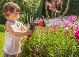 Child hand watering a pink flower garden with hose and spray nozzle