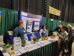 The image shows a booth at a gardening event, hosted by the University of California Cooperative Extension Tulare County Master Gardeners. Several people are interacting at the booth, which is decorated with informational posters, pamphlets, and potted plants. The setting appears lively and educational, with attendees seeking gardening advice and resources.