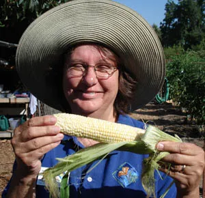 A woman in a wide-brimmed hat holds an ear of corn in a garden.