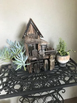 Rustic wooden miniature house sculpture on a wrought iron table, flanked by two potted succulents in gray and terracotta containers.