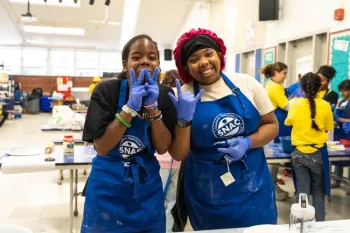 Two girls smiling in a school cafeteria with blue aprons and food prep gloves on.