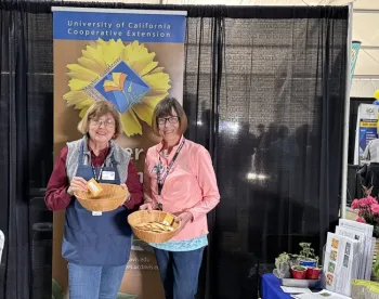 Two volunteers hold baskets of seed packets while standing in front of a Master Gardener banner.