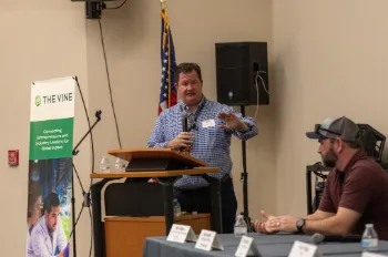 Gabe speaks into a mic toward a table of panel members.