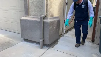 A man spraying the perimeter of a home with a backpack sprayer.