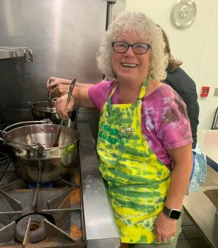 Bobbie Green wears an tie dyed pink shirt and yellow-blue apron while stirring preserves on the stove. 