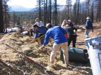 A group of people using hand tools to spread mulch over an eroded area of soil. There are trash cans full of mulch nearby. 