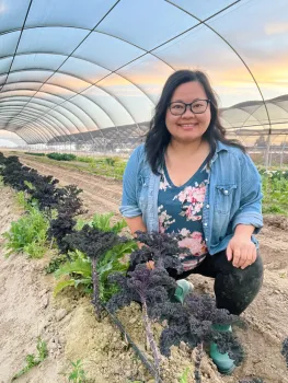 Woman smiling in a blue floral shirt. She is kneeling in a open, uncovered hoop house next to a row of winter kale.