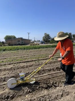 a man wearing an orange shirt and straw hat, pushing a seeder to plant seeds in a field