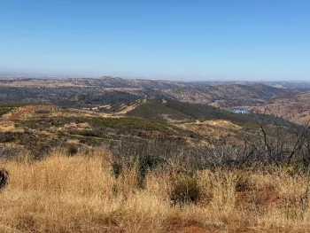 dry brush in front opens to landscape of hills covered by trees and rangelands. A lake is in the valley in the distance