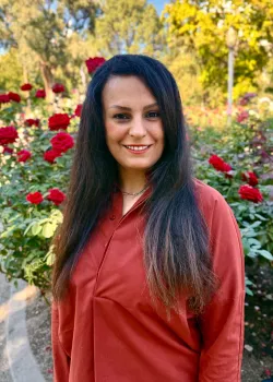 Sara from waist up with red rose bushes behind her
