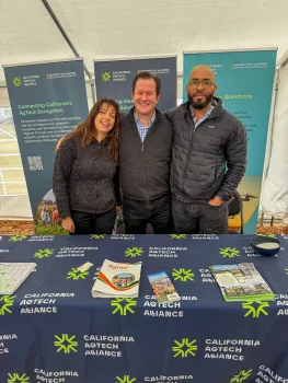 Lucie Cahierre, Gabe Youtsey and Hanif Houston stand at a table with a tablecloth labeled "California AgTech Alliance."