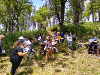 A group of people stand together, holding packets of papers, in an opening in a forest and listen to a speaker present.