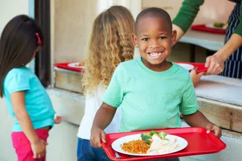 A young boy holds a red tray with a plate of fresh, school food while standing in line at the school cafeteria.