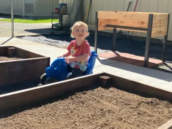 A small child on a blue push tricycle shaped like a fish gazing down into a raised bed, riding the push tricycle in between the raised beds on a path wide enough to accommodate a wheelchair or a push tricycle