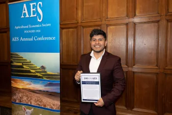 Luis hold a plaque next to a vertical banner that reads: AES Agricultural Economics Society founded 1926. AES Annual Conference