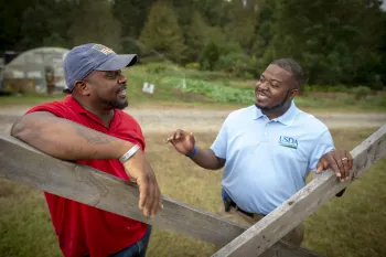 Farmer speaking with a USDA representative with a greenhouse and field in the background.
