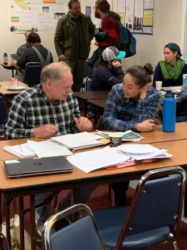 Two people sit at a table and discuss paperwork.