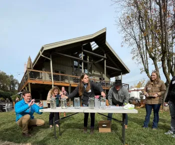 Hope leading a demonstration on sedimentation experiments in glass jars on a folding table, with a blue sky background and a two story house behind with a deck.
