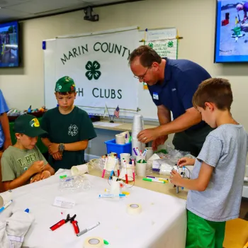 three kids and an adult at a table performing a STEM activity with a Marin County 4-H Clubs banner in the background